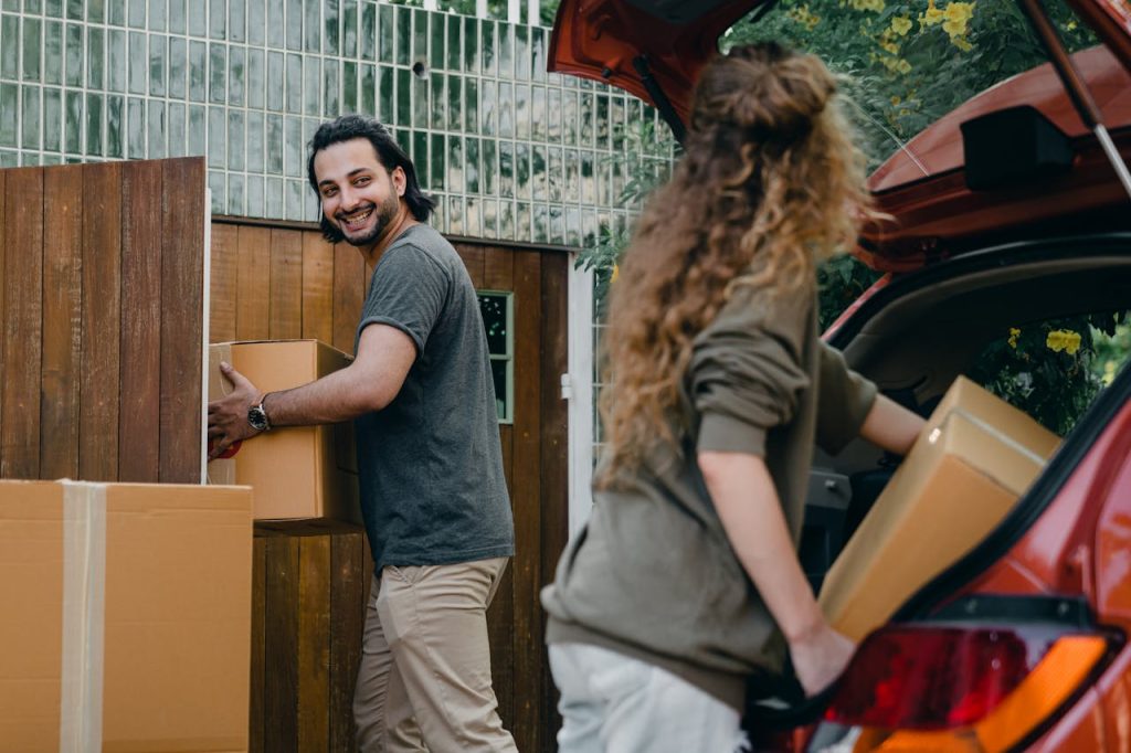 happy-couple-unloading-automobile-while-moving-apartment-in-summer-day-4246266 Cheerful modern male and female in comfortable casual clothes unpacking car trunk in green garden of new house while moving in together