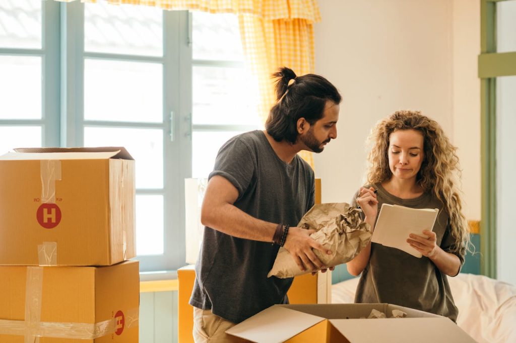 happy-couple-unpacking-boxes-in-new-home-4247768 Cheerful young man and woman smiling while unpacking carton boxes with belongings in new apartment during relocation and looking at paper