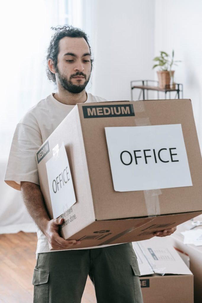 man-in-white-crew-neck-t-shirt-carrying-a-brown-box-7217856 Man moving box labeled Office indoors during relocation or packing.